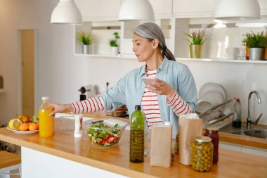 Cep telefonu kullanan ve mutfakta yemek hazırlayan bir kadın.