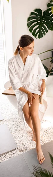 Красивая молодая женщина в халате оттирает ноги натуральной деревянной щеткой, сидя на боку ванны в ванной комнате, украшенной зелеными растениями — стоковое фото