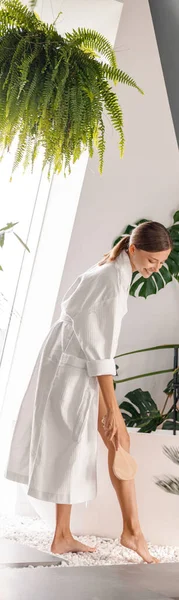 Leende ung kvinna i vit badrock håller träborste för torr borstning ben hud — Stockfoto