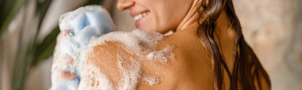Mujer joven feliz aplicando gel de ducha en su cuerpo usando esponja de esponja de esponja mientras toma la ducha —  Fotos de Stock