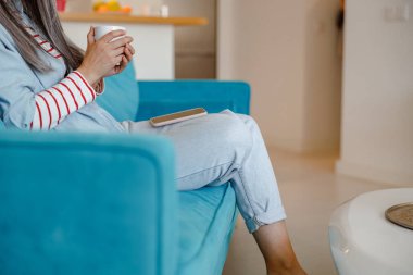 Elinde kahve ve cep telefonu olan bir kadın koltukta oturuyor.