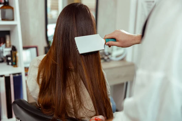 Hand van kapper borstelen lang en glanzend bruin haar van jonge vrouw op schoonheidssalon — Stockfoto