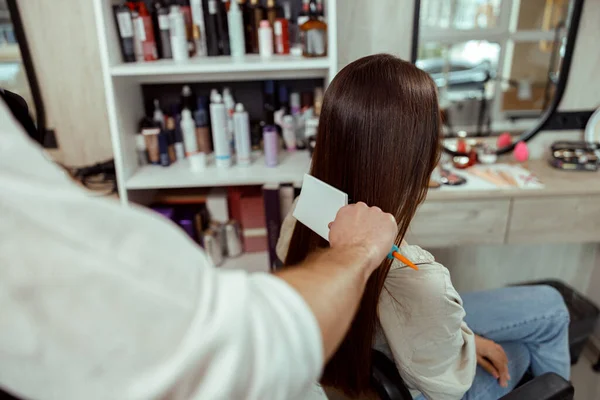 Парикмахер расчесывает длинные и блестящие каштановые волосы молодой женщины в салоне красоты — стоковое фото