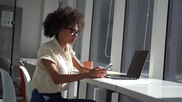 Africano americano sonriente trabajador en moderno minimalista oficina de luz está escribiendo en un teléfono inteligente — Vídeos de Stock