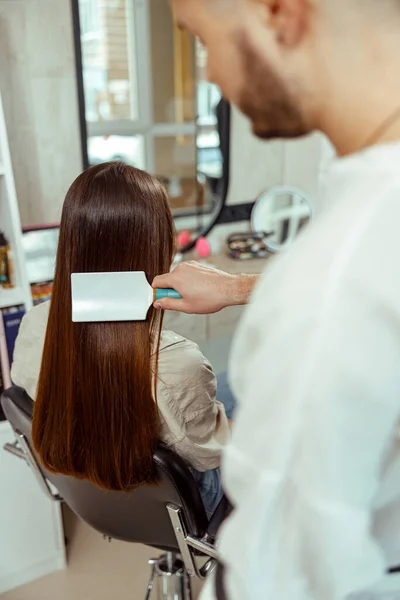 Peluquero cepillado largo y elegante cabello castaño de cliente femenino en el salón de belleza —  Fotos de Stock