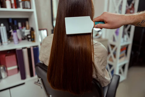 Primer plano del peluquero cepillando el pelo largo y liso marrón del cliente femenino en el salón de belleza —  Fotos de Stock