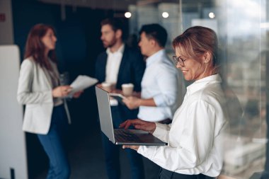 mature confident businesswoman at office with group of colleagues on background, working on laptop
