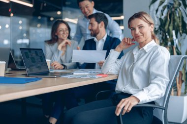 Smiling confident businesswoman sitting on meeting in office with her colleagues at background 