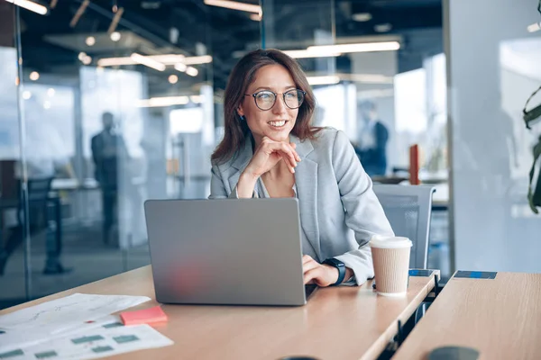 在现代的办公室里，戴着眼镜工作在笔记本电脑上的女商人微笑着，看着窗外的工作环境 — 图库照片