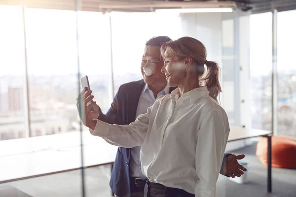 Happy colleagues using smartphone for video call while standing near glass wall in modern office —  Fotos de Stock