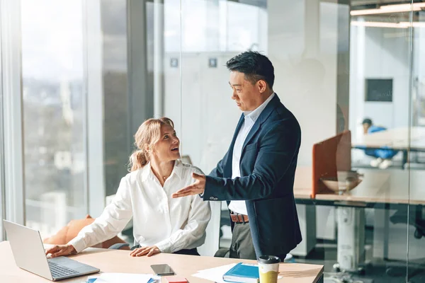Asian businessman is explaining plans to mature caucasian woman that working on laptop in office — Stock Photo, Image
