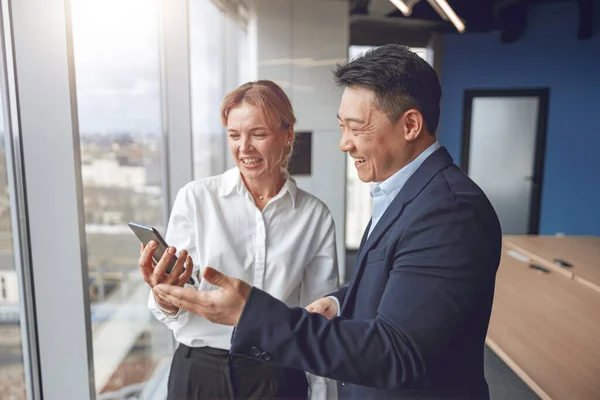 Business workers standing together near window in modern office while using smartphone and smiling — Stock Photo, Image