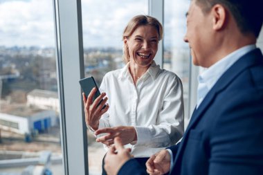 Mutlu iş kadını, elinde akıllı telefon, ofisinde dururken iş arkadaşıyla konuşuyor.