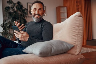 Rahat koltukta otururken cep telefonuyla gülümseyen sakallı adam.