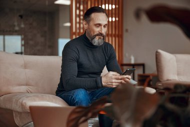 Yetişkin bir adam koltukta oturuyor ve telefon kullanıyor.