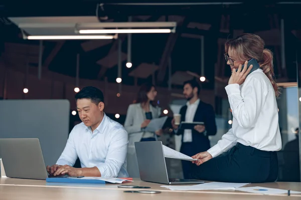 Mature business woman sitting on table and talking phone to solve problem in modern office — ストック写真