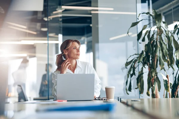 Focused businesswoman working on laptop and looking out the window at workplace at modern office — 스톡 사진