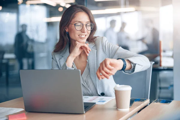 Positive businesswoman wearing eyeglasses looks at her wristwatch at modern office — Stock Photo, Image