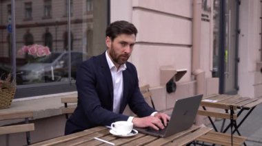 Kafkasyalı yakışıklı işadamı şehirdeki kafenin dışında kameraya gülümsüyor. Laptop klavyesinde mesaj yazılıyor.