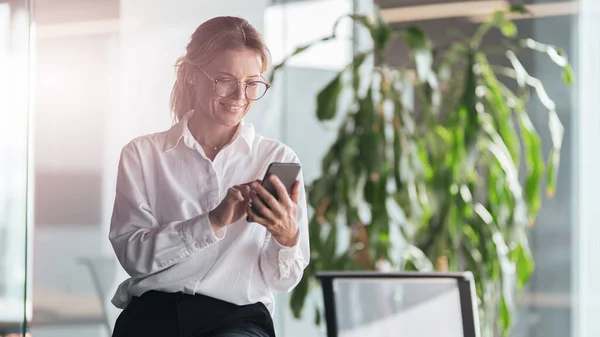 Smiling woman standing in office. Businesswoman with mobile phone in hand looking at smartphone — 스톡 사진