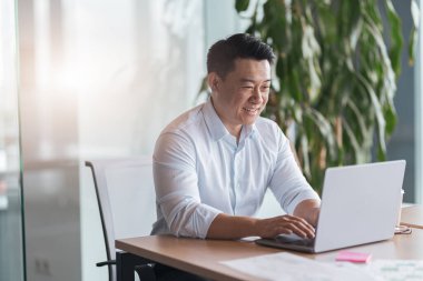 Modern ofisindeki bilgisayarında çalışan gülümseyen Asyalı işadamı.