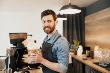 Gülümseyen barista elinde demlikle filtre kahve tutuyor ve kameraya bakıyor.