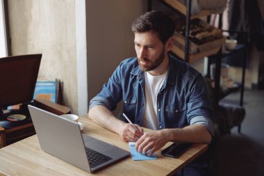 Handsome Caucasian man taking notes in notebook and sitting in cafe at laptop. Freelancer.