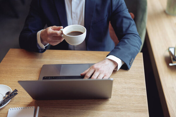 Top view on cup of coffee in male hand, caucasian businessman working at laptop in cafe.