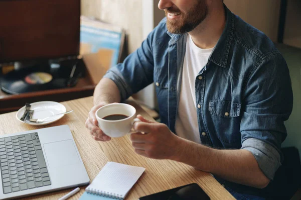 White cup of black coffee in male hand. Caucasian man in cafe working with laptop. Resting.