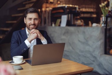 Çekici genç, zarif iş adamı kafede dizüstü bilgisayarda kahve içiyor ve gülümsüyor. Çene ellerde.