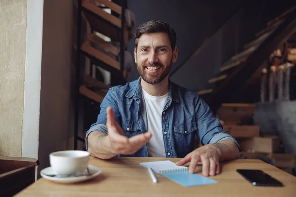 Cheerful attractive man smiling and reaching for camera with hand. Taking gesture. Coffee in cafe.