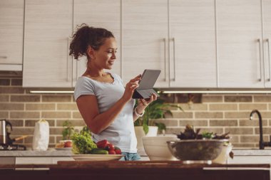 Elinde tabletle güzel bir kadın mutfakta yemek pişirmekle ilgili bloglar yazıyor.