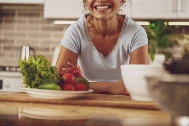 Mutfakta bir tabak sebzeyle masada oturan bir kadının gülümsemesi.
