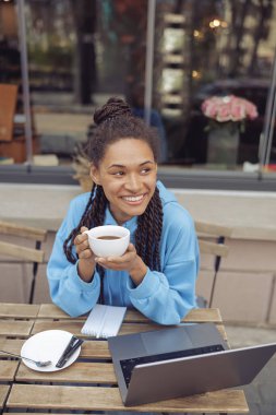 Güzel, genç, melez, stil sahibi bir kadın. Elinde piercing, elinde kahve, gülümsüyor. Üst görünüm.