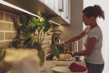 Bir kadın mutfaktaki akan suyun altında, lavabonun üzerinde sebzeleri yıkıyor.
