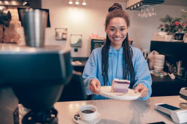 Saçları örgü örüp kameraya cheesecake dağıtan oldukça genç, melez bir bayan barista..