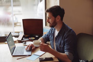 Kafkasyalı yakışıklı erkek, laptopun başında bir fincan kahveyle çalışıyor..