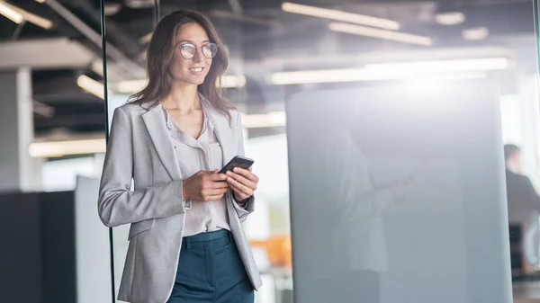 Businesswoman wearing glasses with mobile phone in hand standing in modern office — ストック写真