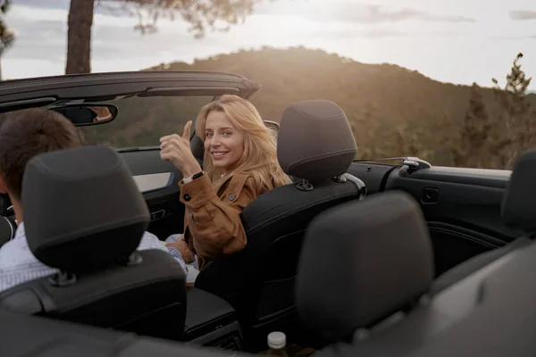 Pretty happy young woman showing thumb up and smiling sitting in cabriolet car with her man — Stock Photo, Image