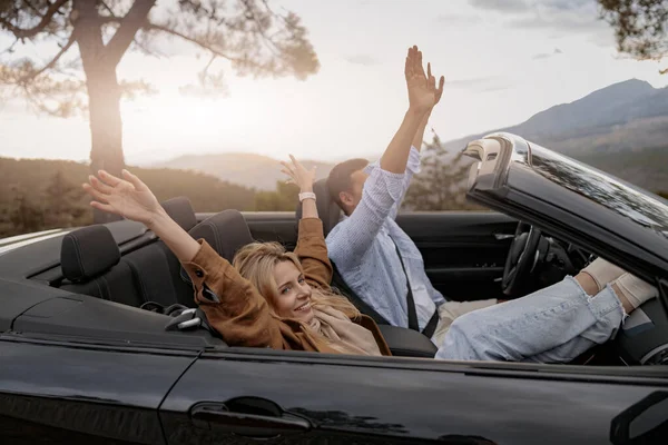 Side view of cheerful boyfriend and girlfriend sitting in convertible car in mountains — Stock Photo, Image