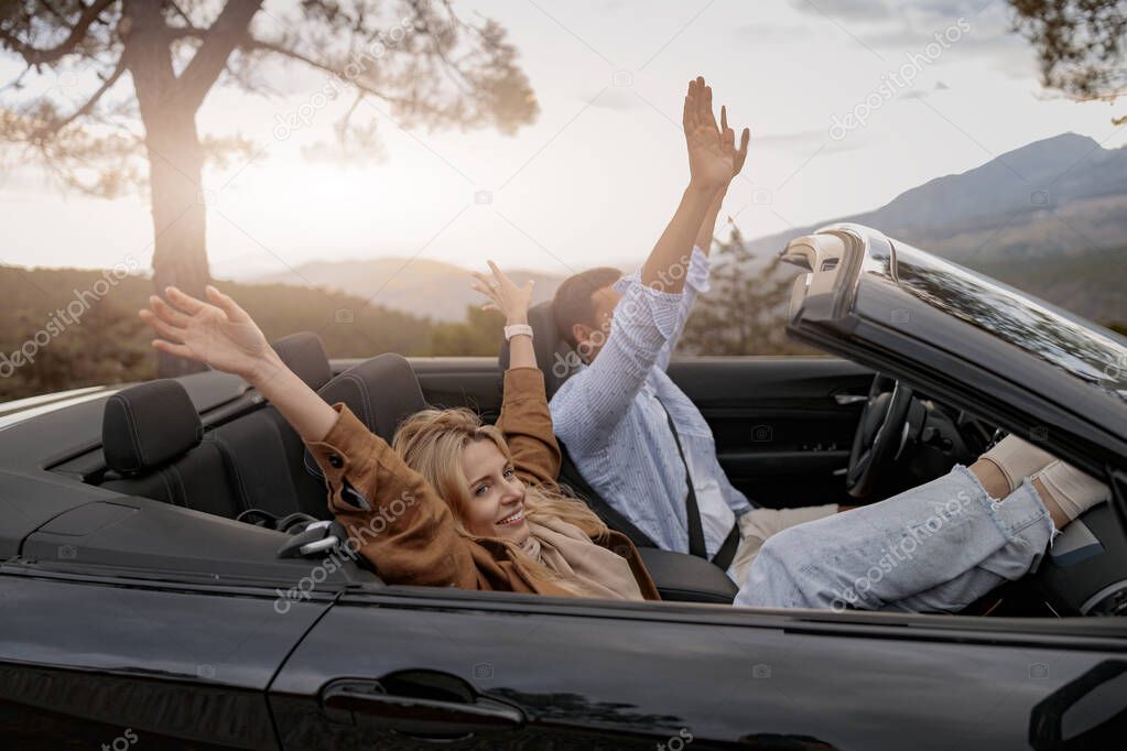 Side view of cheerful boyfriend and girlfriend sitting in convertible car in mountains