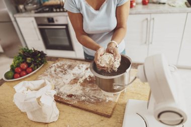 Mutfakta bir kadın hamur yoğurucunun hamurunu alıp tahtaya yuvarlıyor.