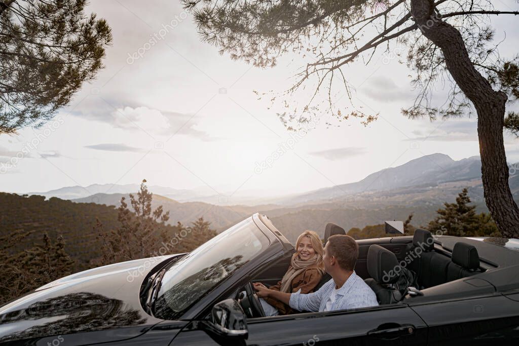 Young joyful family couple on a trip driving in cabriolet convertible car between mountains