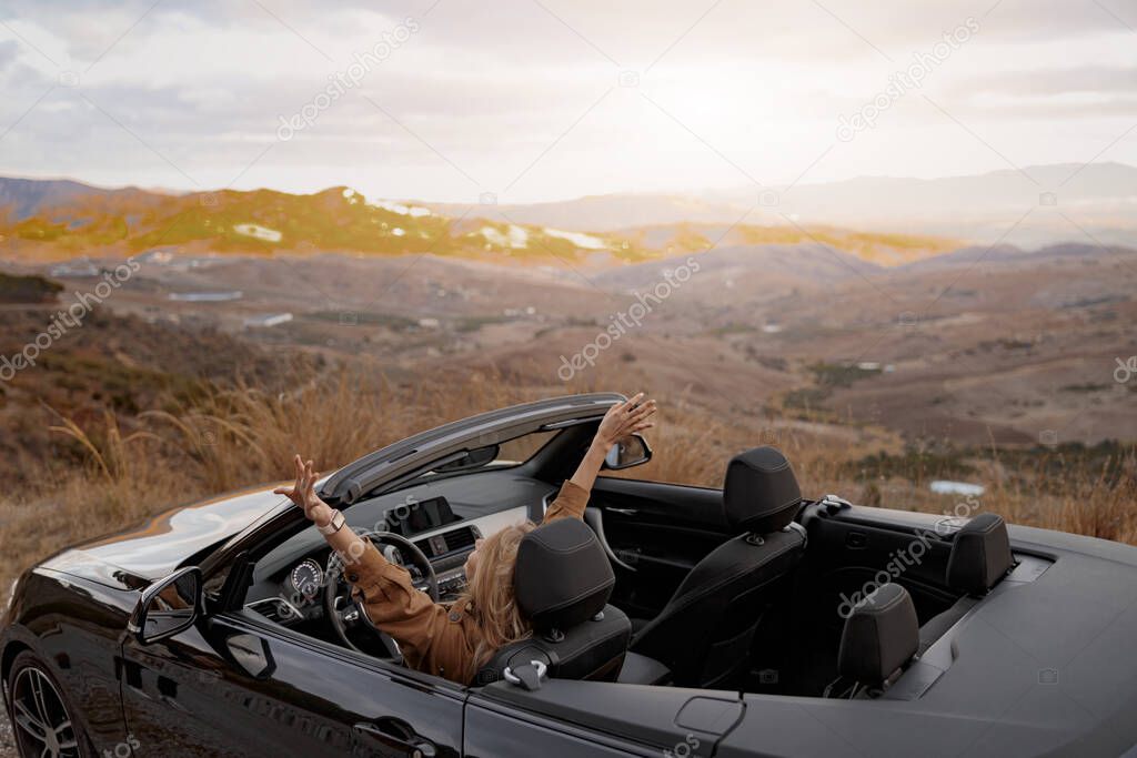 Rear of young happy female holding hand up feeling free sitting in convertible car by mountainside