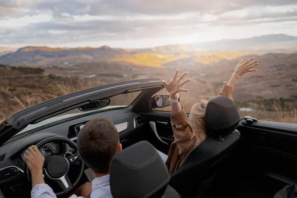 Rear of couple man and woman traveling in convertible car by mountainside enjoying panorama view