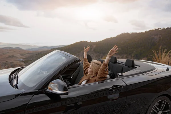 Young happy smiling female traveling in convertible car by mountainside holding hands up, side view