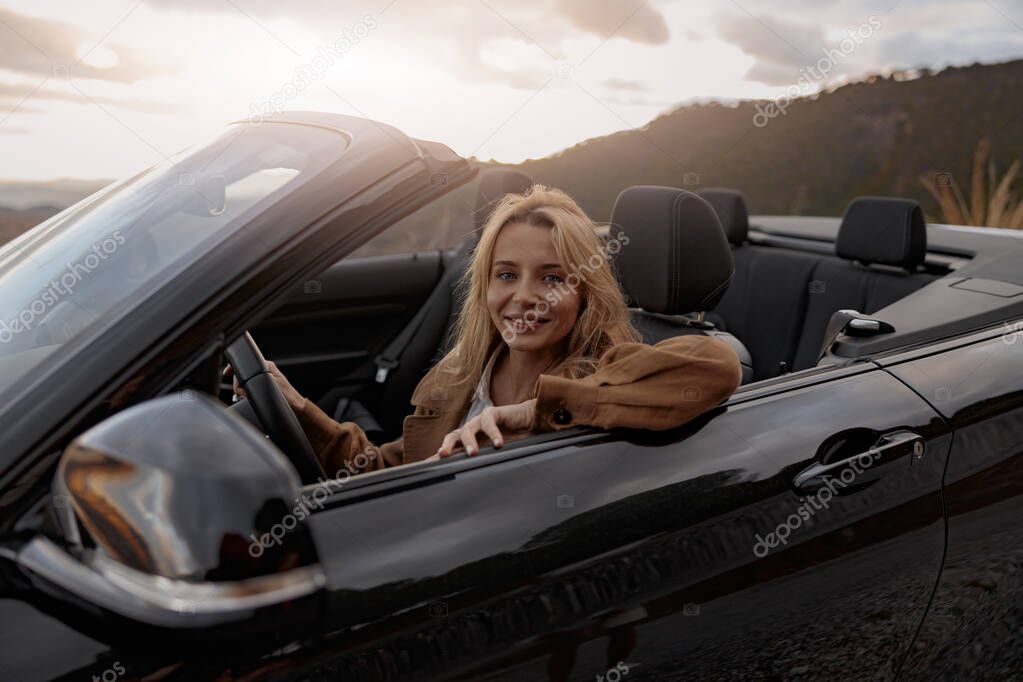 Close up of joyful young pretty blonde woman smiling to camera sitting in convertible car at sunset
