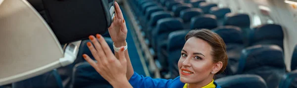 旅行スーツケースを頭上の手荷物室に置きながら航空機の旅客サロンに立つ笑顔の女性客室乗務員 — ストック写真