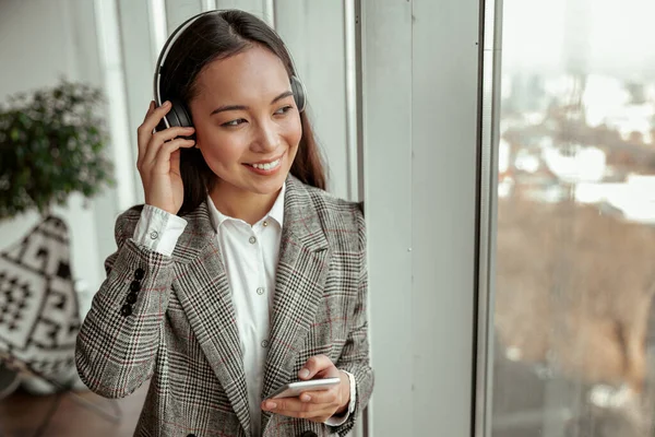 Asian Business woman listening music from phone in headphones standing near window in office