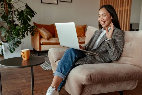 Gülümseyen Asyalı iş kadını müşteriye video çağrısı yapıyor ve el sallıyor rahat ofisinde oturuyor.